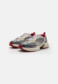 Sport sneakers featuring a cream, grey, and red mesh upper, with a thick white midsole and textured rubber outsole. Accented with logo details.