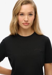 Zwart katoenen T-shirt met korte mouwen, met een ronde halslijn en een klein reliëf "Superdry" logo op de borst. Glad textuur.