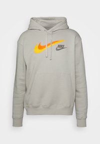 Ljusgrå luvtröja med en framficka, med en djärv orange och gul Nike-logotyp och en mindre grå textlogotyp på bröstet.