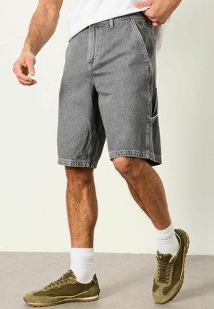 Mann trägt graue Jeansshorts, weiße Socken, olivgrüne Sneaker und ein weißes Hemd, steht vor einem schlichten hellen Hintergrund.