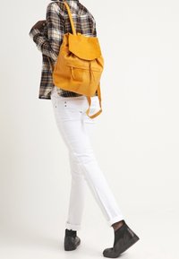 Mochila amarilla con textura suave, cierre superior plegable y cordón, combinada con pantalones blancos y zapatos negros, sobre un fondo neutro.