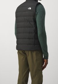 Man som bär en svart vadderad väst från North Face, en grön långärmad skjorta och olivgröna byxor, sedd bakifrån mot en neutral bakgrund.