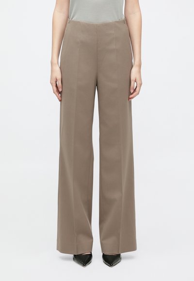 Weite Hosen aus einem muted braunen Stoff mit glatter Textur, hoher Taille und Bügelfalten. Kombiniert mit spitzen schwarzen Schuhen.