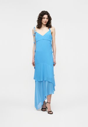 Femeie cu păr creț purtând o rochie asimetrică albastru deschis fără mâneci, cu stratificare de țesături, și pantofi negri cu toc și deschiși în față, pe un fundal alb.