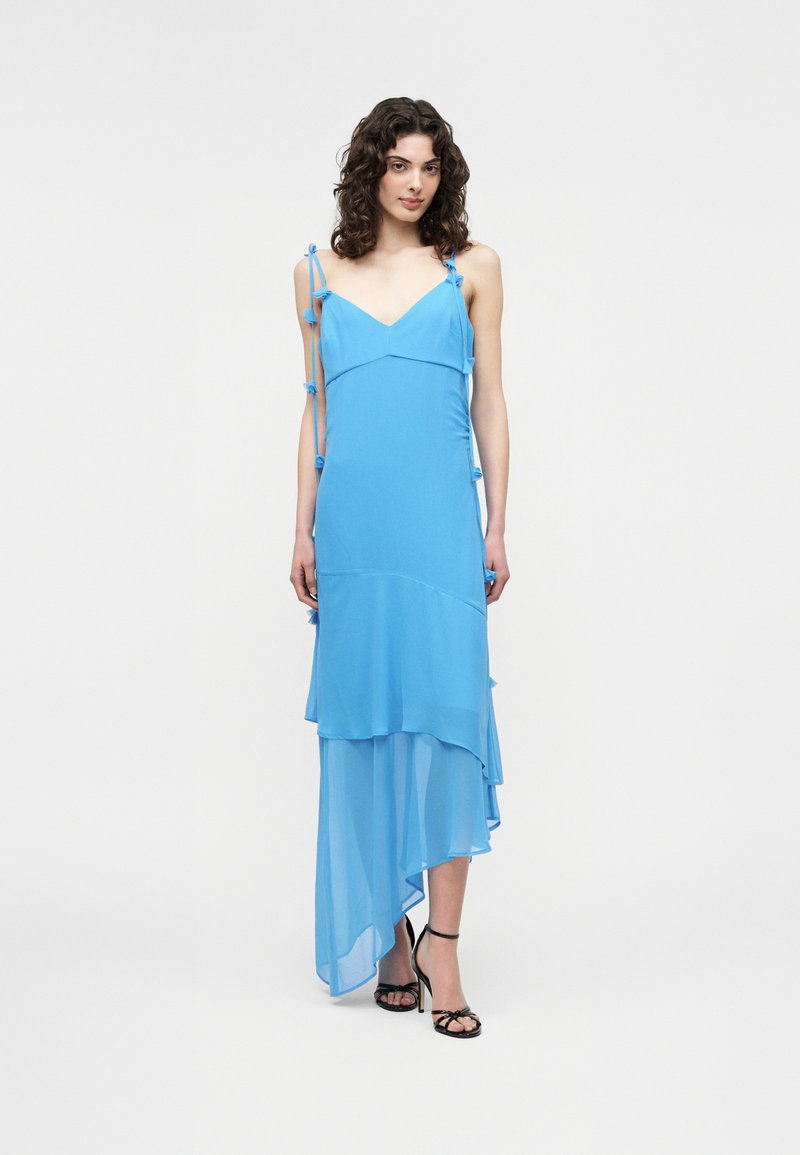 Γυναίκα με σγουρά μαλλιά που φοράει αμάνικο ασύμμετρο φόρεμα σε ανοιχτό μπλε χρώμα με στρωματοποιημένο ύφασμα και μαύρα πέδιλα με ανοικτά δάχτυλα, σε λευκό φόντο.