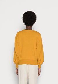 Person med kort lockigt hår som bär en mörkgul långärmad tröja och ljusbeige byxor, stående med ryggen mot en ljus bakgrund.