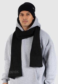 Black knitted scarf and beanie set, featuring ribbed texture and a logo detail. The scarf has a flat-ended design, worn over a gray hoodie.