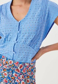 Blusa blu a occhielli con bottoni e dettagli in pizzo verticale abbinata a una gonna floreale nei toni del rosa, arancione e blu, mano poggiata sull'anca.