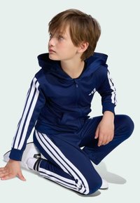 Chlapec klečící a dívající se nahoru, oblečený v tmavě modrém teplákovém kompletu s bílými pruhy a bílých teniskách na světlém pozadí.