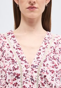 Γυναίκα που φοράει ροζ και βουργουνδί μπλούζα με λαιμόκοψη V και δαντελένιο τελείωμα, ασημένια σκουλαρίκια κρίκους και ίσια καστανά μαλλιά.