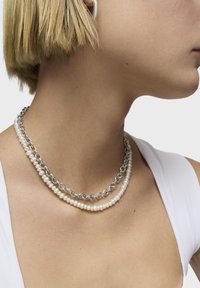 Collana a strati con maglie di catena d'argento e perle bianche, che mostra un mix di texture e forme su uno sfondo color pelle.