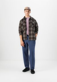 Jonge man in een beige pet, met een bruine geruite overhemd over een paarse t-shirt, blauwe broek en zwarte sneakers, staande tegen een witte achtergrond.
