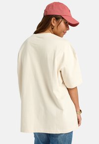 Cream-colored oversized t-shirt with short sleeves, paired with a pink baseball cap. The model's hair is in loose waves, with gold hoop earrings.