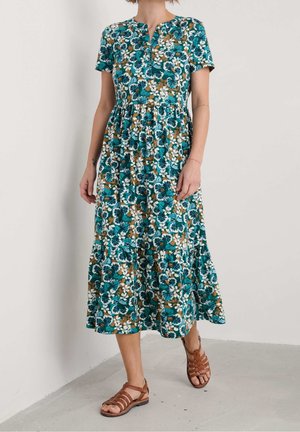 Femme portant une robe midi à manches courtes à fleurs de couleur bleu sarcelle, marron et blanc, avec des sandales plates marron à lanières, marchant à l'intérieur sur un sol gris.
