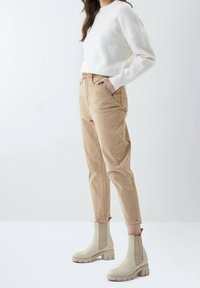 Pantalones de talle alto beige, textura suave, corte casual. Suéter blanco con puños acanalados. Botines de tobillo beige claro con suela gruesa y diseño slip-on.