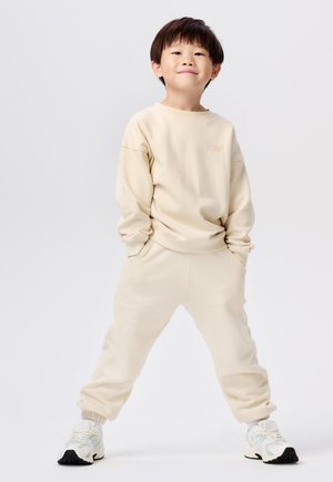 Beige katoenen sweatshirt en bijpassende joggingbroek, relaxte pasvorm, geribbelde manchetten, met geborduurd logo op de borst, gecombineerd met witte sneakers.