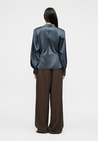 Γυναίκα με μακριά σκούρα μαλλιά που φοράει μπλούζα από μπλε σατέν και ευρύχωρο καφέ παντελόνι, στέκεται γυρισμένη προς τα πίσω σε απλό φόντο.