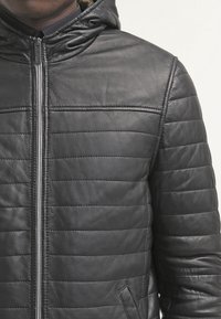 Giubbotto in pelle nera con cappuccio, texture trapuntata, cuciture verticali e chiusura con zip, dotato di tasche laterali.