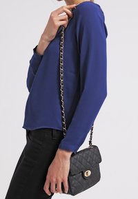 Sort quiltet håndtaske med en guld kædedyse, båret som crossbody. Blå langærmet bluse, tætsiddende sorte bukser, set fra siden.