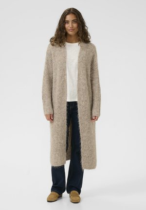 Beige langer Cardigan aus weichem, flauschigem Material, mit offener Vorderseite und geripptem Textur, kombiniert mit einem hellen Oberteil und dunklen Jeans.