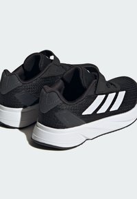 Zapatillas deportivas negras con parte superior de malla, diseño de tres rayas blancas y detalles en gris. Presentan una suela acolchada y un ajuste seguro.