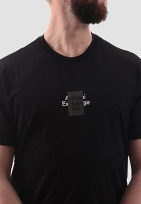 La camiseta de algodón negra presenta un parche central con el texto "Armani Exchange 1991" en blanco sobre un fondo negro. Diseño clásico de cuello redondo.