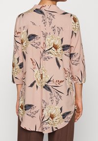 Ljusrosa blus med blommönster i beige och marinblått. Har en krage, trekvartsärmar och en rundad nederkant. Mjuk tygtextur.