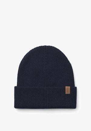 Hombre con barba que lleva un gorro de punto azul marino y una chaqueta de forro polar marrón claro con una cremallera oscura sobre un fondo blanco.