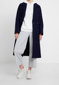 Marineblauer Peacoat mit doppelt geknöpftem Verschluss über einem wei ßen Hoodie, kombiniert mit hellen karierten, gekürzten Hosen und weißen Sneakers.