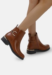 Botas de tobillo de imitación de cuero marrón con una textura suave, cremallera lateral, acento dorado en el tacón y un tacón bajo tipo bloque.