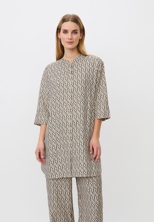 Vrouw staand, gekleed in een losse, langemouwige, patroonrijke tuniek en bijpassende wijde broek, tegen een eenvoudige witte achtergrond.