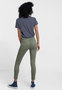 Pantalon skinny vert olive avec poches arrière, associé à un t-shirt rayé bleu et blanc. La silhouette porte des baskets montantes bleues.