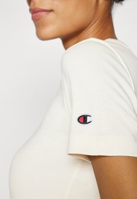 Lysecreme bomulds t-shirt med fitted design, korte ærmer og et broderet logo i sort, rød og hvid på ærmet.
