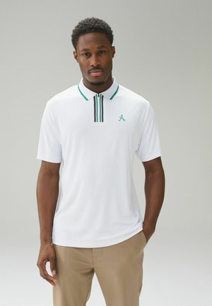 Mann trägt weißes Poloshirt mit grünem Rand und beige Hose, steht vor einfachem hellgrauem Hintergrund.