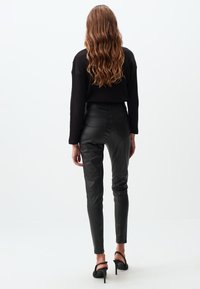 Pantaloni aderenti neri simili alla pelle con vita alta, abbinati a un top nero morbido a costine, mostrati di spalle. Tacchi neri con cinturini.