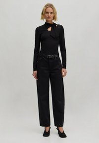 Schwarzes langärmeliges Oberteil mit einem Cut-Out-Detail am Hals, kombiniert mit hochgeschnittenen, weiten schwarzen Hosen und schwarzen, hohen Schuhen.