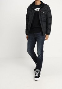 Černá prošívaná bunda s límcem z umělé ovčí vlny, nošená přes černou mikinu, v kombinaci s modrými džíny a černými teniskami s bílými akcenty.