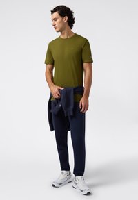 Man in een olijfgroene T-shirt, marineblauwe joggingsbroek, witte sneakers, met een marineblauwe sweatshirt om zijn middel geknoopt, staande tegen een effen achtergrond.