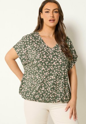 Vrouw met lang bruin haar draagt een groene blouse met korte mouwen met kleine roze en witte bloemen, gecombineerd met lichtgekleurde broek.