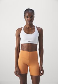 Sujetador deportivo blanco con tirantes anchos y logotipo en el centro; pantalones cortos naranja de cintura alta con costuras inclinadas y textura suave.