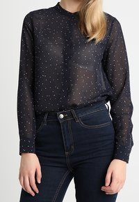 Mörkblå transparent blus med vita prickar, knäppning fram, bandad krage och lång ärm, kombinerad med mörka denimjeans.