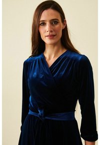 Samettiin kääritty sininen mekko, jossa on kolme neljännesosaa hihoista, solmittava vyö vyötäröllä ja V-kaula-aukko. Sujuva pinnan rakenne ja tyylikäs laskos.