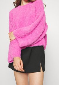 Fluffig rosa tröja med långa ärmar och rund halsringning; matchad med en svart kjol med en sidospalt. Mjuk textur och livlig färg.