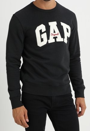 Mann trägt ein schwarzes Sweatshirt mit großen weißen "GAP"-Buchstaben und kleinem rotem "Original"-Schriftzug auf der Brust, kombiniert mit schwarzen Hosen.