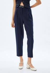 Navyblauwe op maat gemaakte broek van gladde stof; heeft een voorpiek, riemlussen en een ontspannen pasvorm; gecombineerd met witte blokhakken.