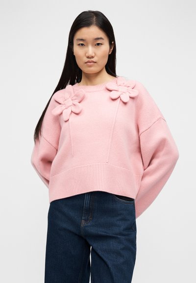 Ροζ πλεκτό πουλόβερ με μεγάλα λουλουδένια εφαρμοστικά στα μανίκια, ριμπ τελείωμα και χαλαρή γραμμή. Συνδυάζεται με σκούρο μπλε τζιν.