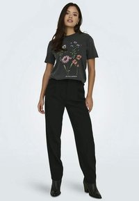 Noor naine kannab musta lilledega graafilist T-särki, musti kõrge vöökohtadega pükse ja musti pahkluu saapaid, seistes ühtlase tausta ees.