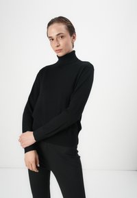 Black turtleneck sweater made of soft fabric, featuring long sleeves, ribbed cuffs, and a relaxed fit. Worn with black fitted pants.