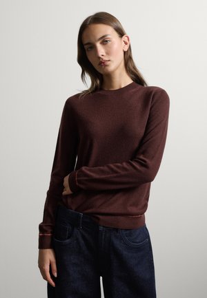 Giovane donna con capelli castani lisci indossa un maglione marrone a maniche lunghe e jeans blu scuro, in piedi davanti a uno sfondo chiaro e semplice.
