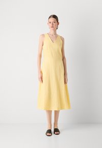 Rochie galbenă din in, cu decolteu în V, fără mâneci, cu un corp ajustat și fustă A-line fluidă. Pur dată cu sandale negre tip slide și bijuterii simple.